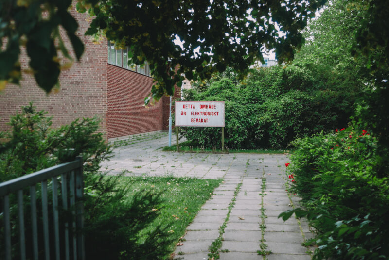 A brick house, some bushes and a sign with the text "Detta område är elektroniskt bevakat".